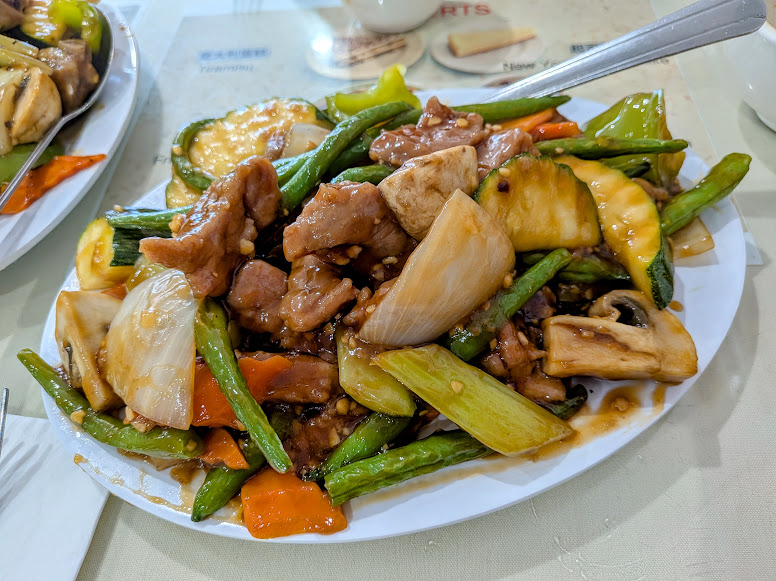 Plate of stir-fried vegetables and meat, including green beans, carrots, zucchini, mushrooms, and onions, covered in a brown sauce with a metal spoon.