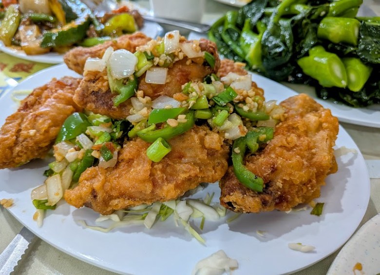 Plate of fried fish topped with chopped onions and green peppers, served on a bed of shredded cabbage, with a side of cooked leafy greens in the background.