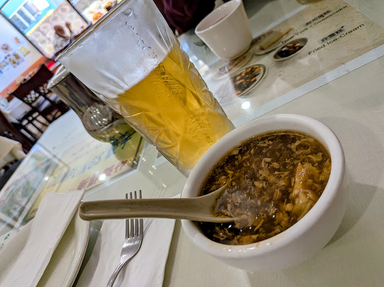 A bowl of hot and sour soup with a ceramic spoon next to a glass of iced tea on a restaurant table.