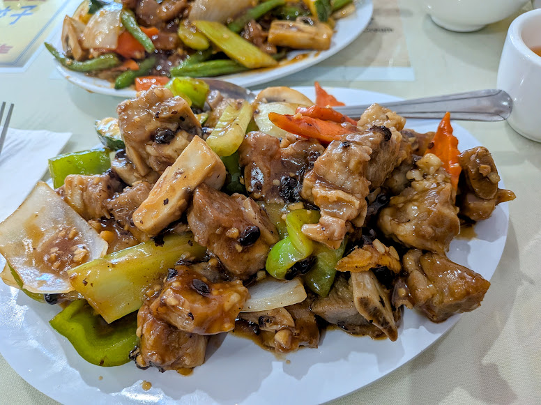 Plate of stir-fried pork ribs with black bean sauce, bell peppers, onions, and carrots, served on a white dish with a spoon and fork on the side.