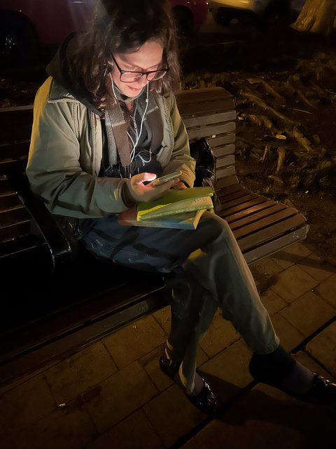A person sits on a bench at night, looking at their phone while holding an open book in their lap, wearing glasses, earphones, and a jacket.