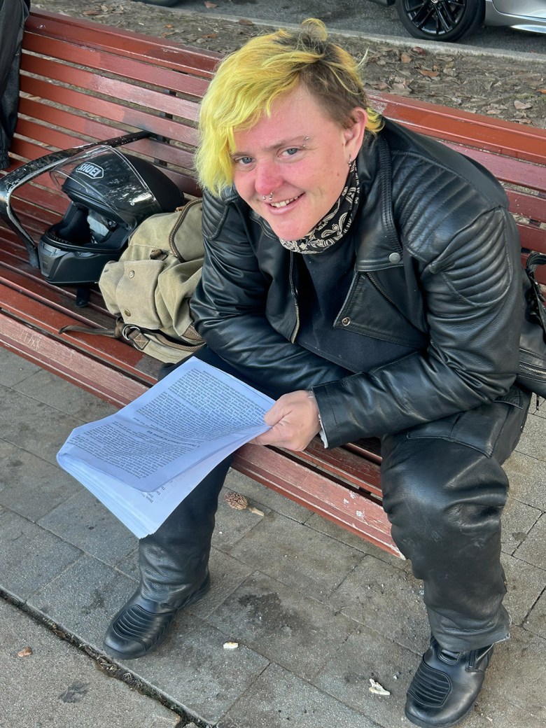 Person with short yellow hair, black leather jacket, and boots sits on a bench holding papers. A helmet and bag are beside them.