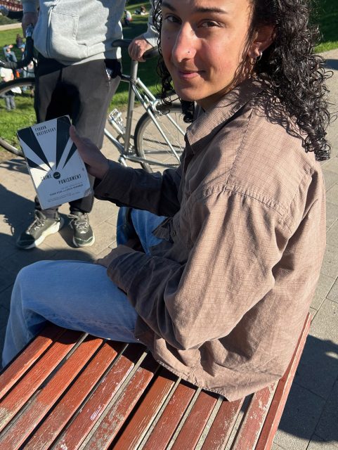 Person with long curly hair sits on a bench outdoors, holding a black-and-white book. A bicycle and another person are visible in the background.