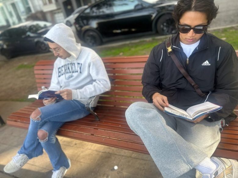 Two people sit on a wooden park bench reading books; one wears a grey Berkeley hoodie, the other wears a black Adidas jacket and sunglasses.
