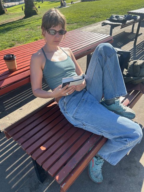 A person wearing sunglasses reads a book while lounging on a red park bench. A coffee cup and backpack are on a nearby bench.