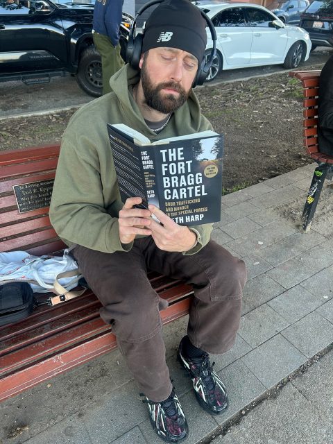 A man wearing headphones and a beanie sits on a bench reading “The Fort Bragg Cartel” book, with cars parked in the background and a backpack beside him.
