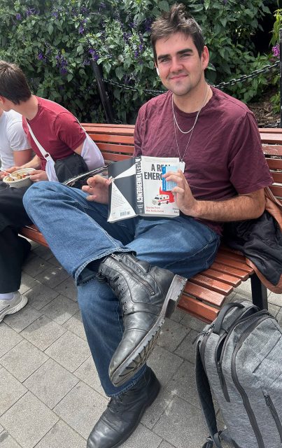 A person in a maroon shirt and jeans sits on a bench, holding a book and notebook, with a gray backpack on the ground and greenery in the background.