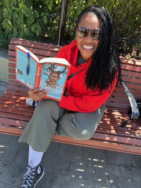 Person in sunglasses and a red hoodie sits on a bench outdoors, smiling and reading a book titled “Imagination” by Nic Stone and Angie Thomas.