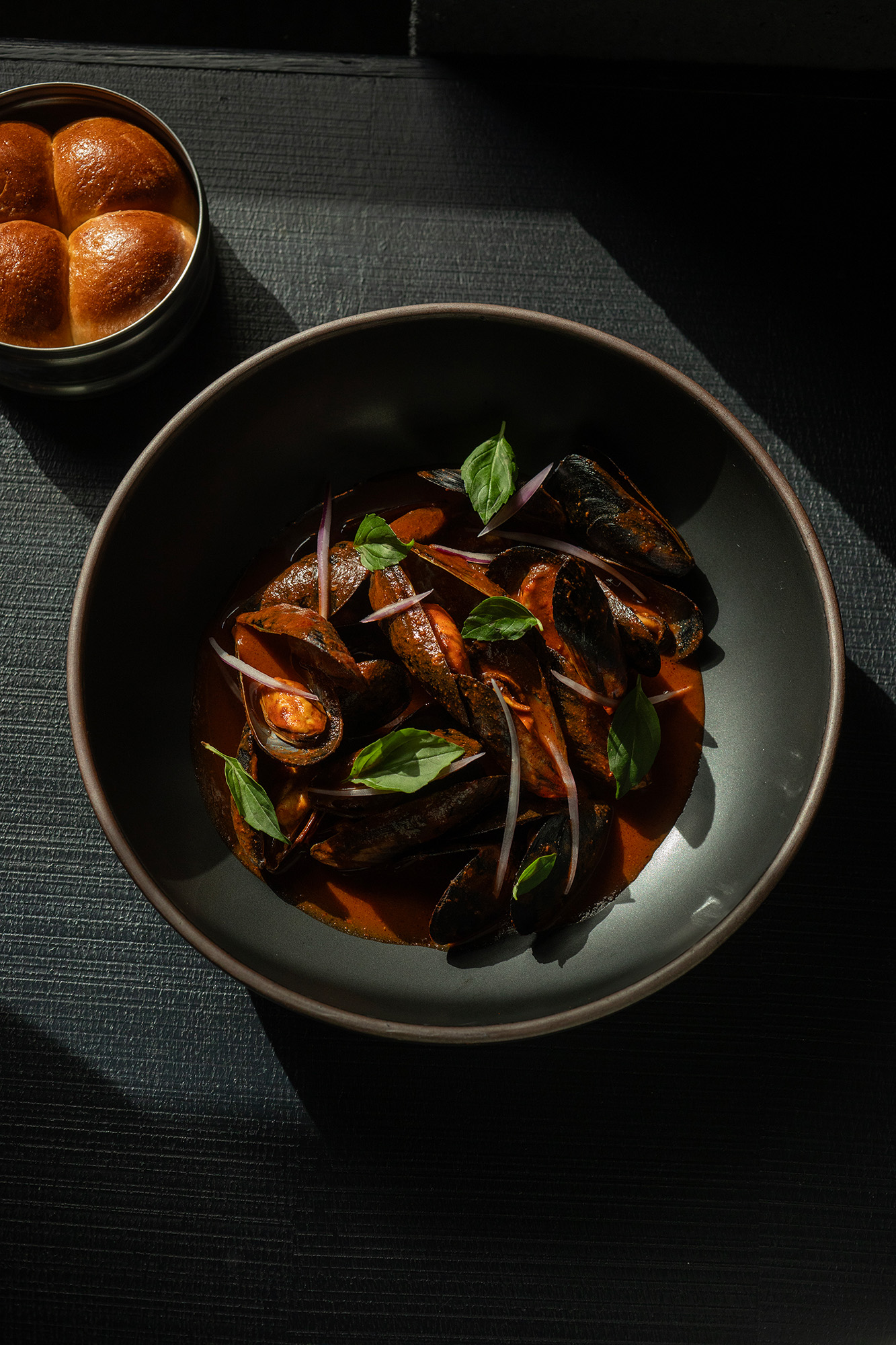 A dish of mussels in a red sauce, with rolls of bread on the side.
