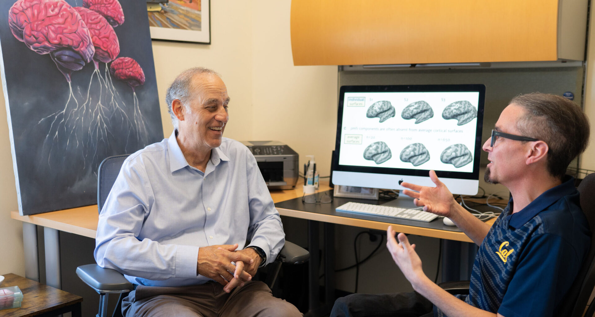 two men seated and talking in front of computer monitor with brains on the screen