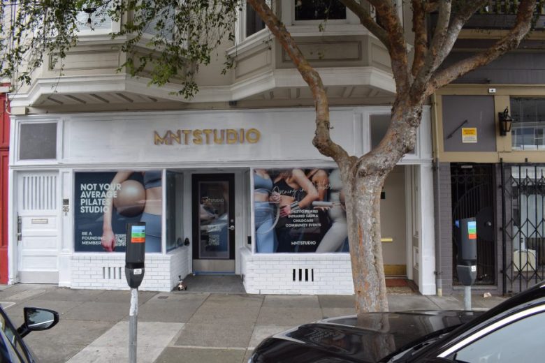 Street view of MNTSTUDIO pilates studio entrance, with large front windows displaying signage and photos, and parking meters along the sidewalk.
