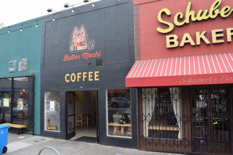 Storefronts of Butter Mochi Coffee and Schubert’s Bakery side by side; a person sits inside Butter Mochi Coffee near the window, and the sidewalk and street are visible outside.