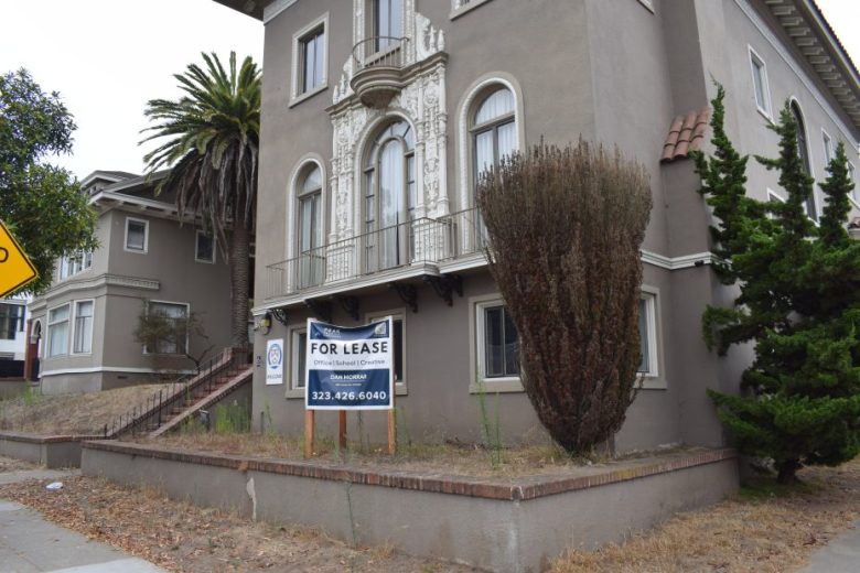 A beige two-story apartment building with a “For Lease” sign in front, overgrown bushes, and a palm tree on the left side.