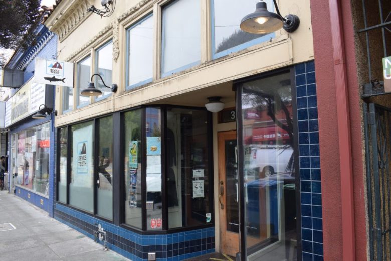 Street view of a beige and blue tiled storefront with large windows, glass door, and signage above. Adjacent businesses and sidewalk are also visible.