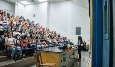 A photograph of a large, brightly lit university lecture hall. A female instructor stands at the front, facing a large group of students seated in tiered rows. She is speaking and gesturing slightly with her hands. At the front of the room is a large wooden demonstration desk. The students are facing the instructor, with some taking notes or using laptops. Another person sits in a chair in the background.