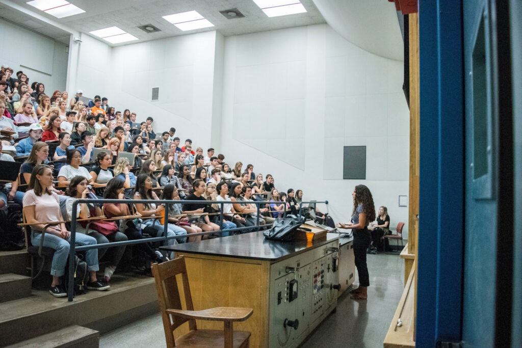 A photograph of a large, brightly lit university lecture hall. A female instructor stands at the front, facing a large group of students seated in tiered rows. She is speaking and gesturing slightly with her hands. At the front of the room is a large wooden demonstration desk. The students are facing the instructor, with some taking notes or using laptops. Another person sits in a chair in the background.