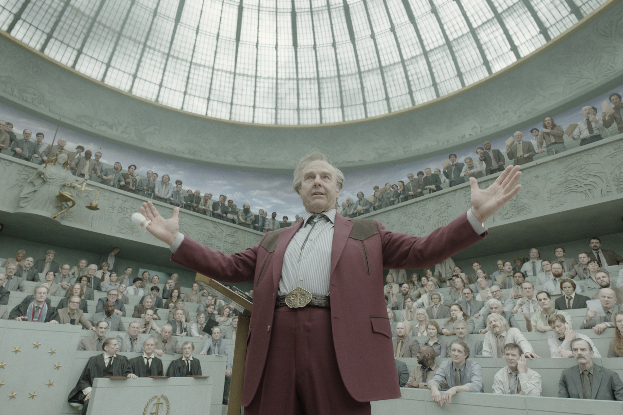 older white man raises arms in auditorium with large crowd watching