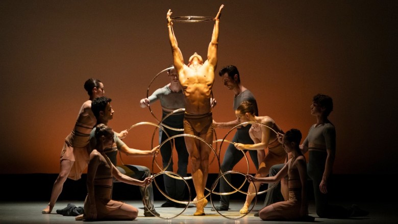 A male dancer holds a ring above his head, surrounded by five other dancers in muted costumes, all performing on a softly lit stage with an orange background.