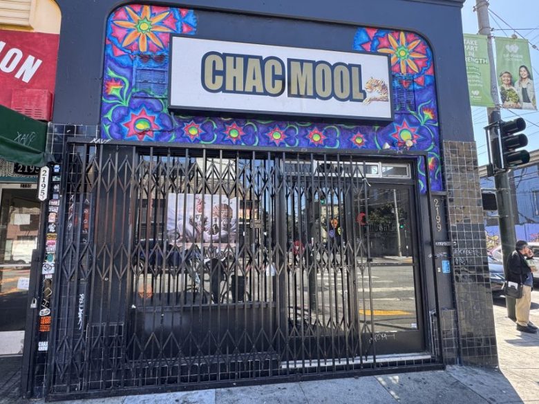 Storefront with a metal gate reads “CHAC MOOL” on a white sign above, framed by colorful mural art; street and sidewalk visible in front.