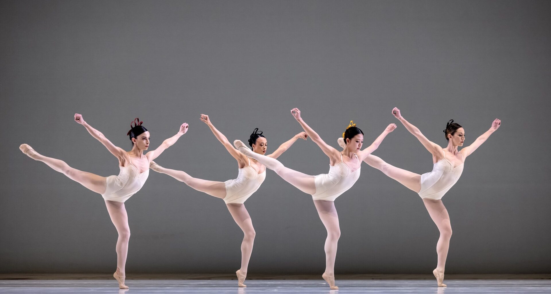 Four female dancers in white leotards, white tights, and white slippers do an arabesque facing stage left in a staggered line. They bend their supporting knee, pressing over their standing foot, and hodl their arms up in a V shape, fists clenched.