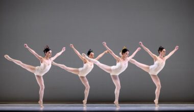 Four female dancers in white leotards, white tights, and white slippers do an arabesque facing stage left in a staggered line. They bend their supporting knee, pressing over their standing foot, and hodl their arms up in a V shape, fists clenched.