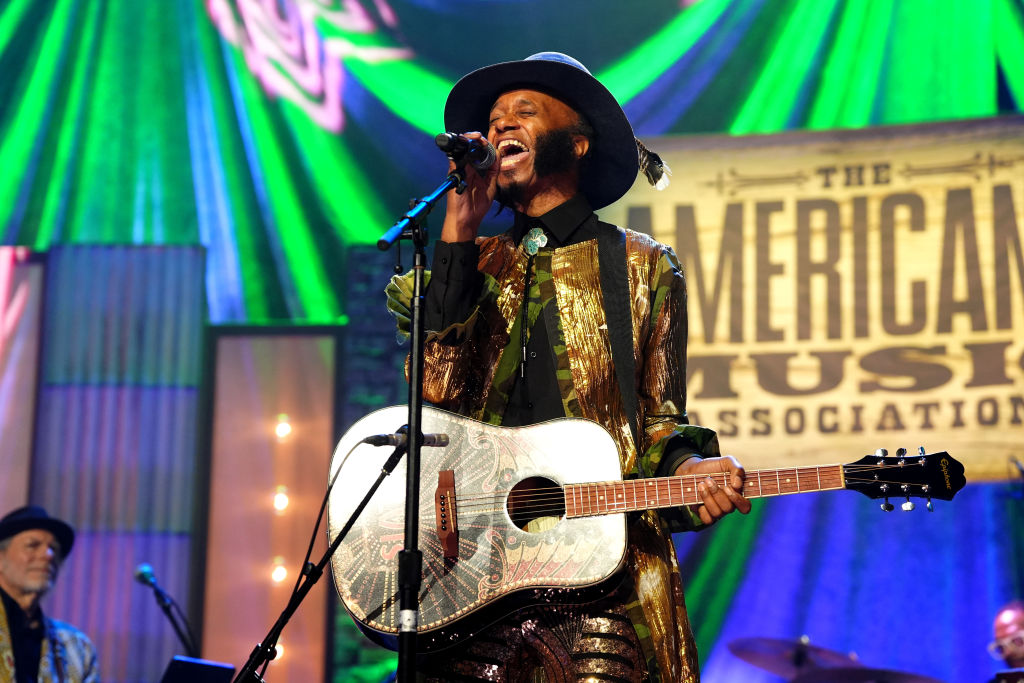 a Black man with a guitar, gold outfit and hat sings into a microphone onstage while holding a guitar