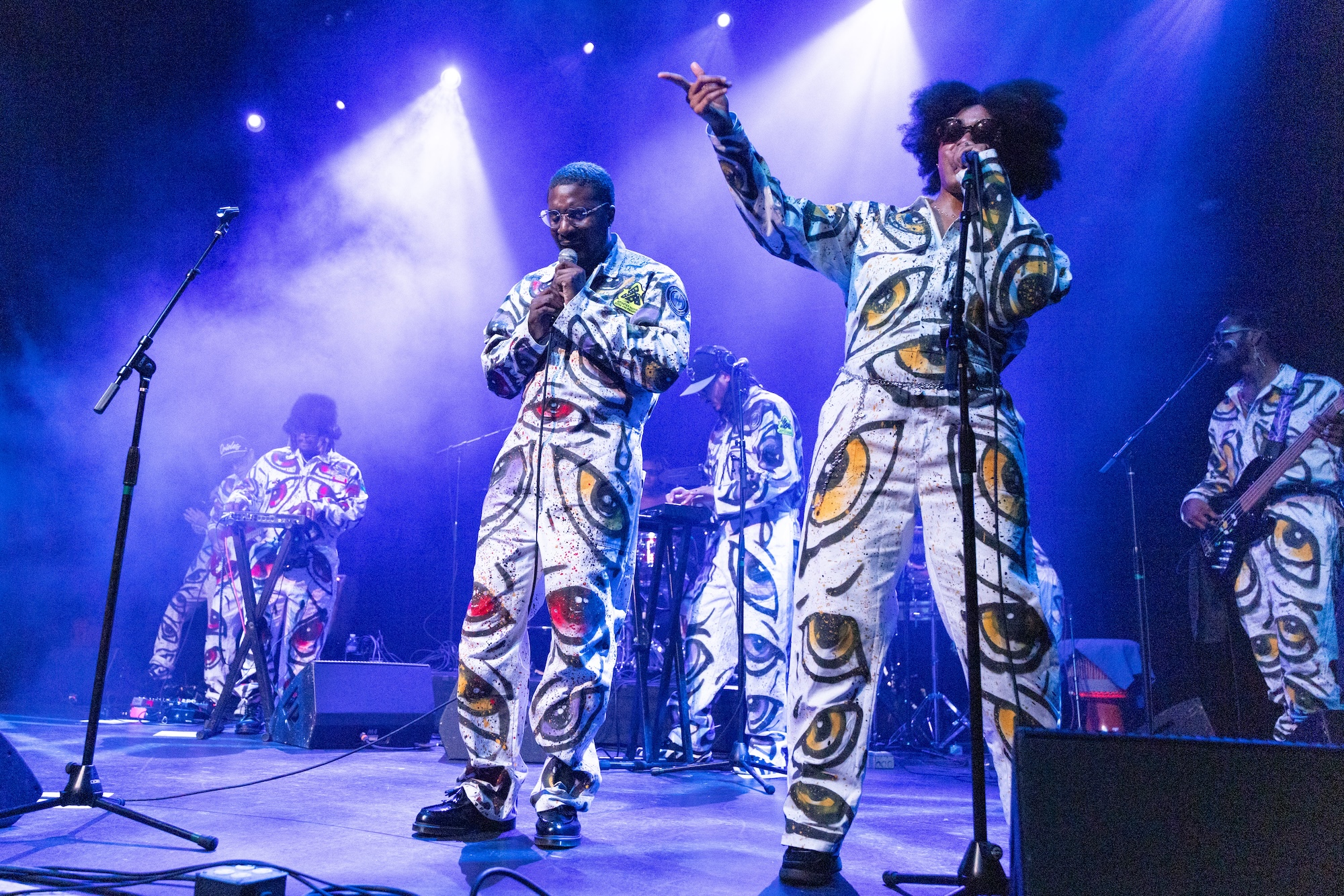 A Black hip-hop artist raps into a microphone, flanked by backup singers and musicians. 