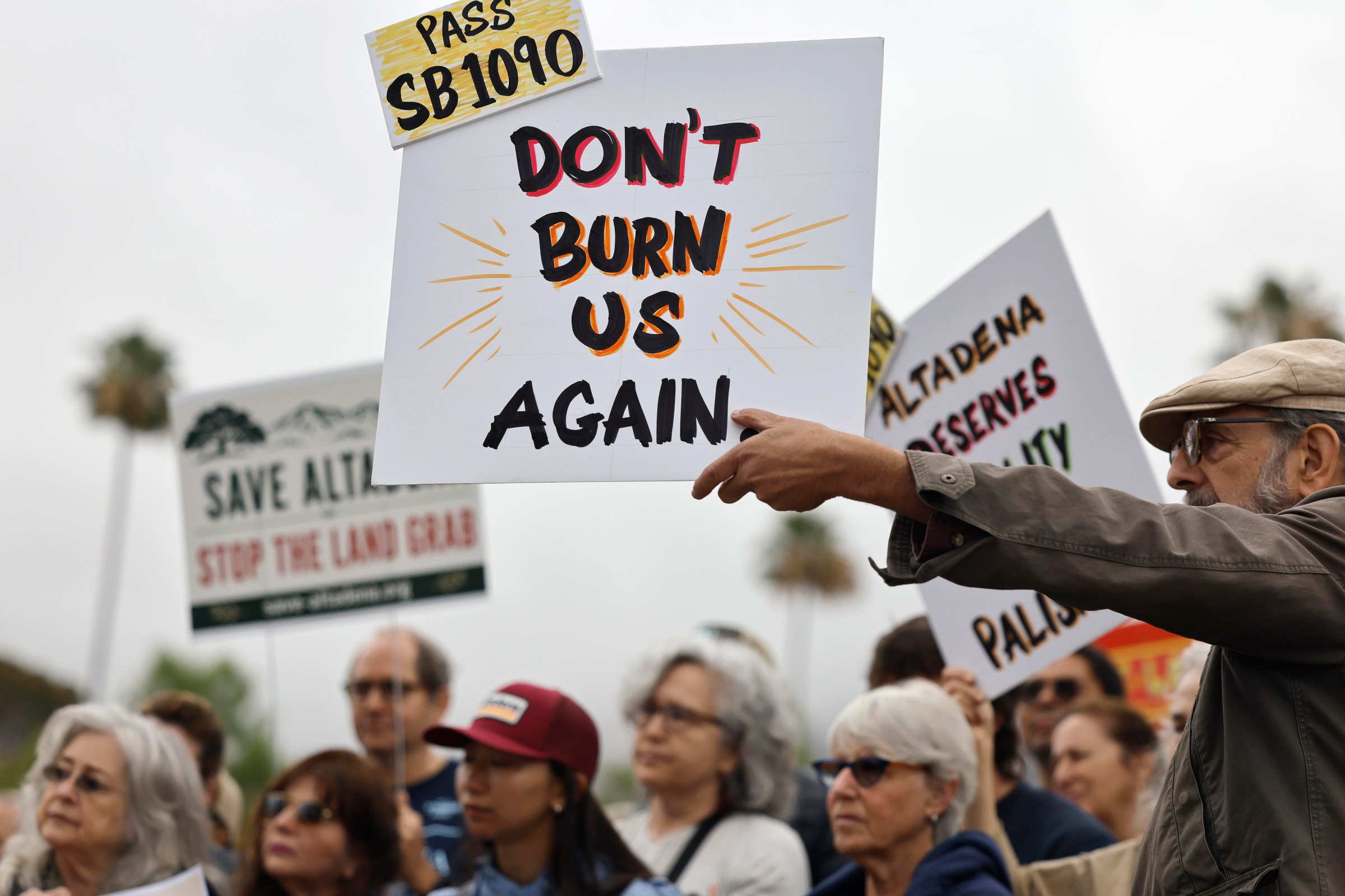 Protesters in Altadena hold signs supporting SB 1090, including one reading “Don’t burn us again,” during a demonstration over post-fire rebuilding and housing policy.