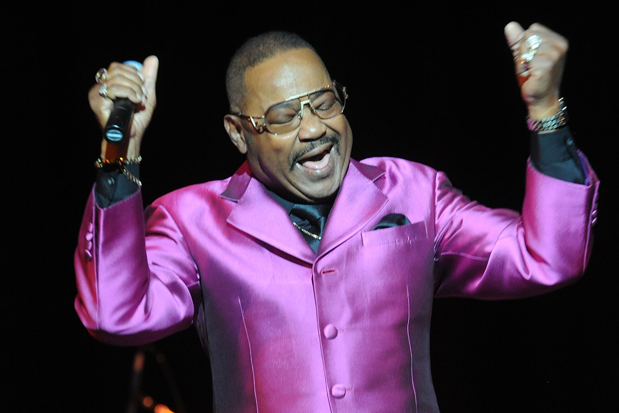 An older Black man in a shiny purple suit jacket smiles with eyes shut and arms raised in the air