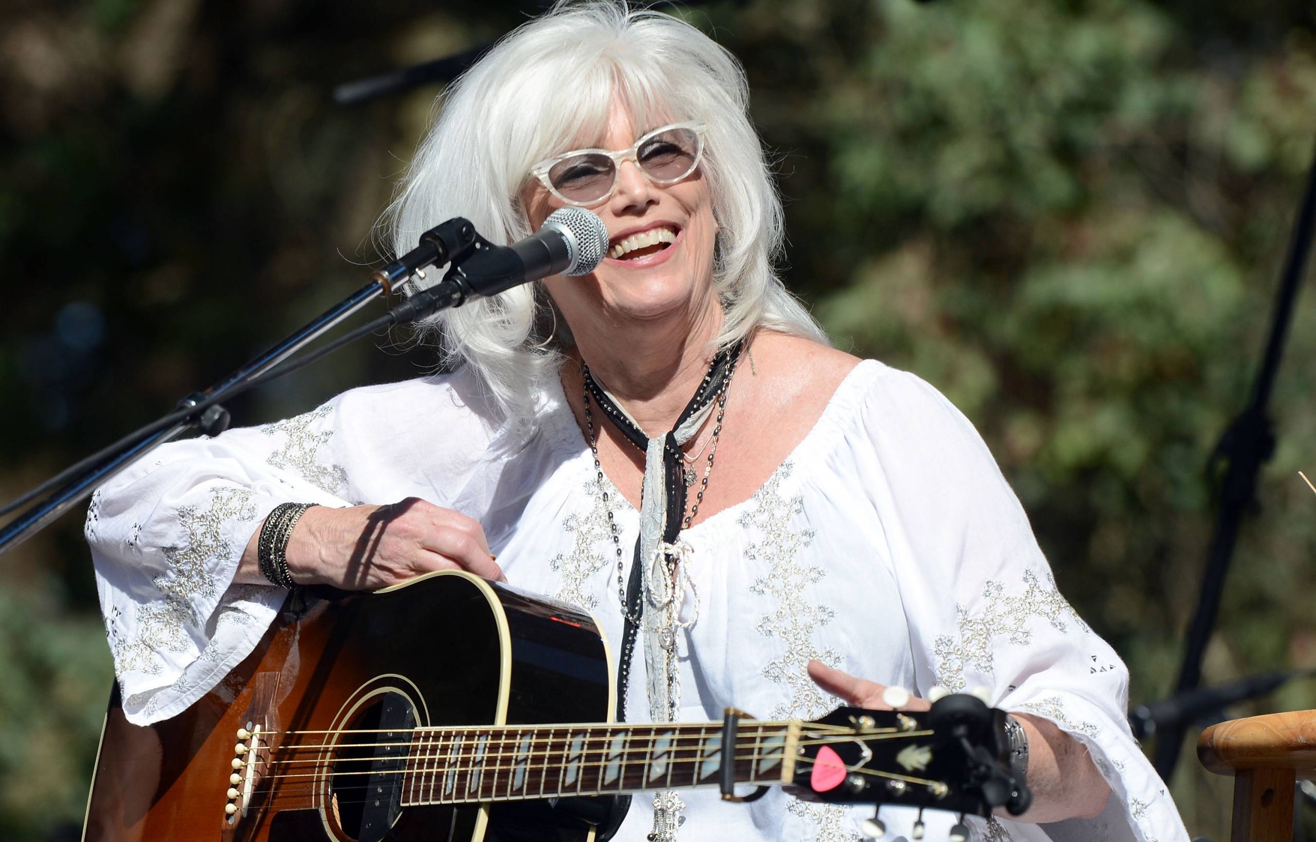 a woman with white hair and sunglasses smiles as she plays guitar on an outdoor stage