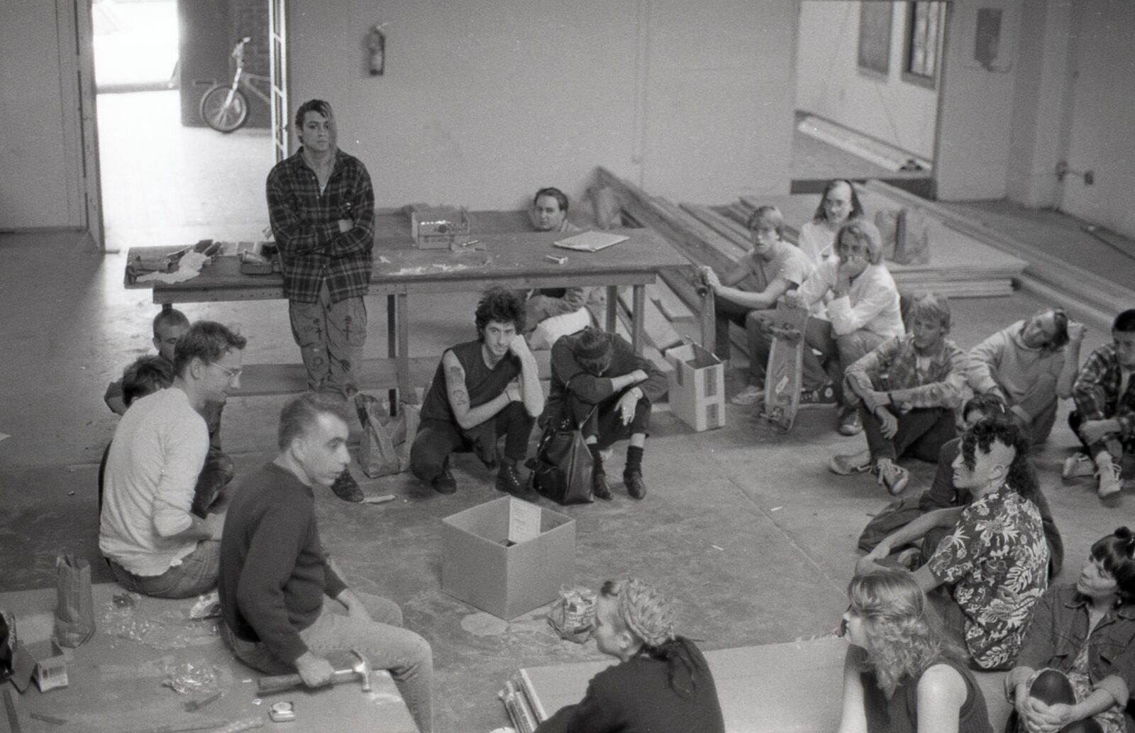A group of people sit in a circle in a building that looks like an active worksite, with lumber and tools scattered around. Their attention is focused on an older man holding a hammer.