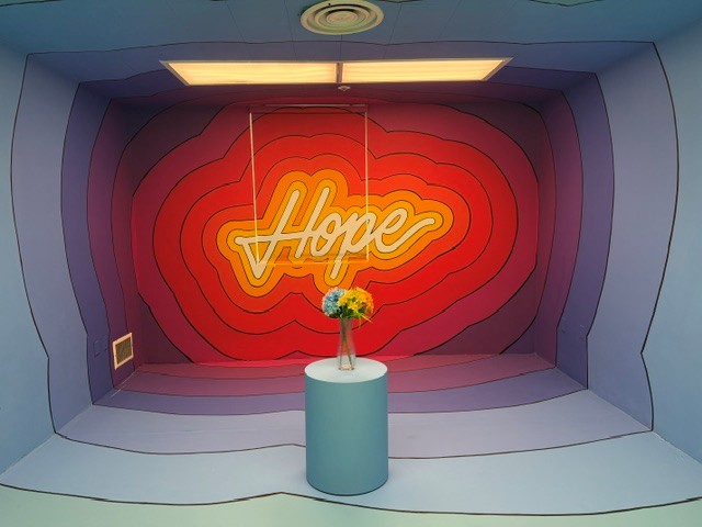 Neon "Hope" sign glowing inside a colorful immersive room with a pedestal holding fresh flowers at Hospital of Emotions.