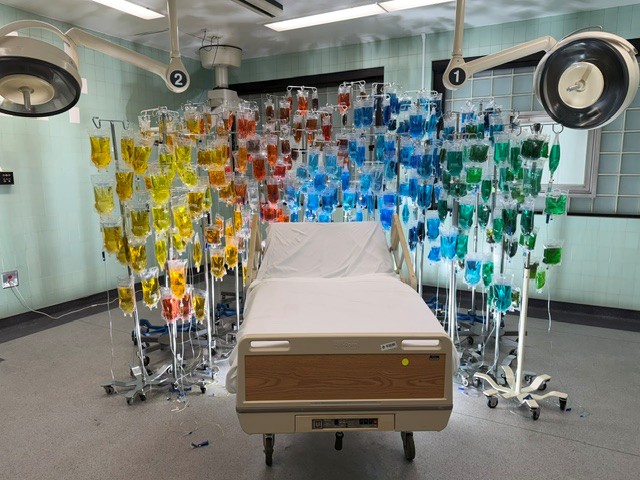Hospital bed surrounded by rows of colorful hanging IV bags in a former operating room art installation.