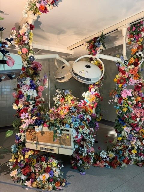Hospital bed engulfed by cascading flowers beneath surgical lights in a former operating room transformed into an immersive art installation.