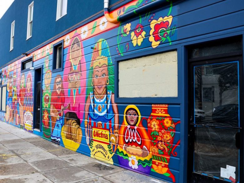 A colorful mural featuring traditional Mexican motifs, including dolls, flowers, and pottery, decorates the side of a blue building along a sidewalk.