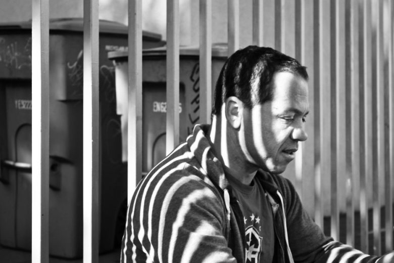 A man sits near vertical metal bars, with shadows from the bars creating striped patterns on his face and clothing. Two large trash bins are visible in the background.