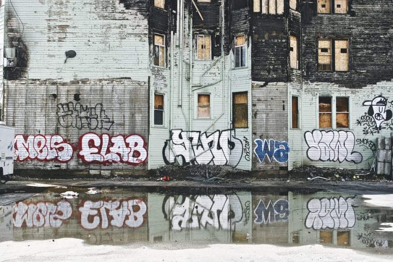 Graffiti-covered, boarded-up building with peeling paint is reflected in a large puddle on the ground in front of it.