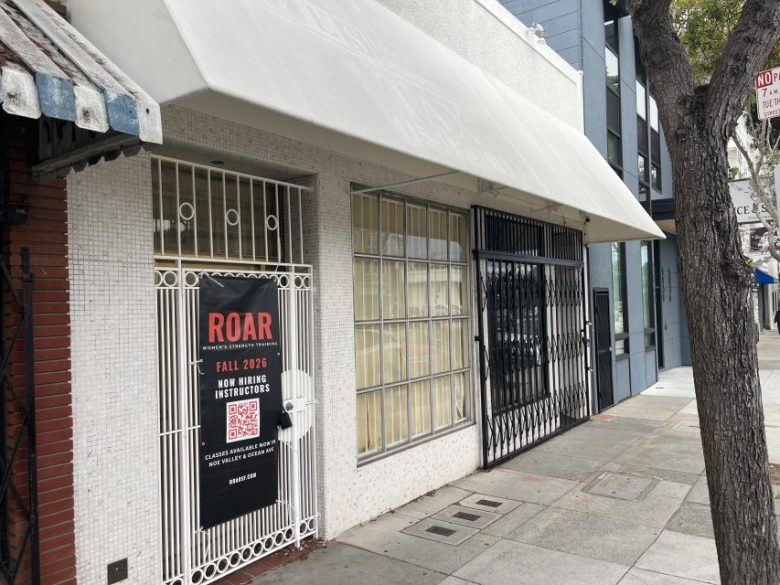 A white storefront with metal gates displays a ROAR sign recruiting fall 2026 instructors. The sidewalk, a tree, and other commercial buildings are visible.