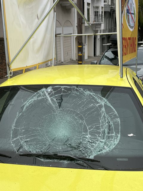 A yellow car with a large, circular crack in the center of its windshield is parked on the street near a sign and other vehicles.