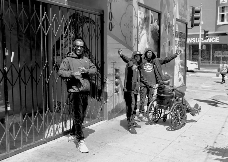 Four people are on a city sidewalk; one uses a wheelchair, and the others stand nearby. A storefront and street signs are visible.