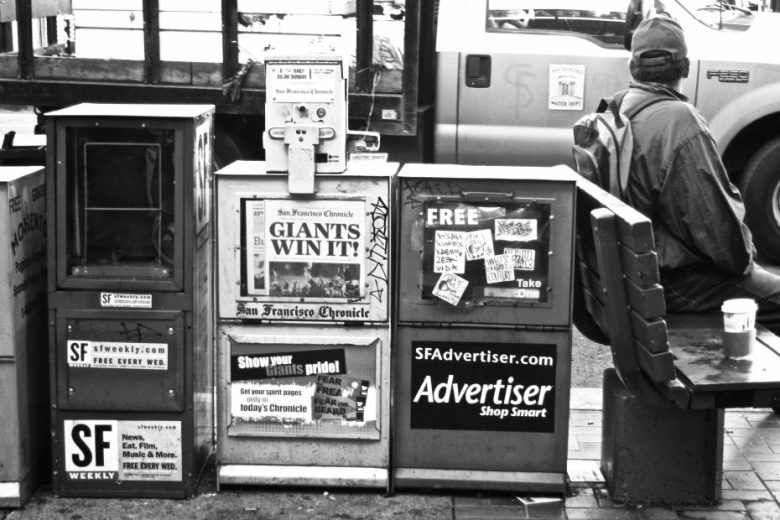 Three newspaper vending machines and a bench with a seated person wearing a hat and backpack are on a city sidewalk; a van is visible in the background.