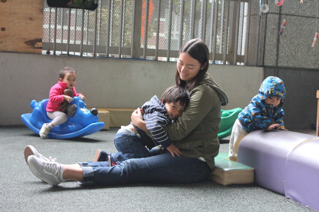 An adult sits cross-legged on a gray cushioned floor in a playroom, giving a warm hug to a smiling toddler boy wearing a navy striped hooded jacket and jeans. To the left in the background, a child wearing a bright pink jacket sits on a blue plastic rocking dog toy. To the right, another toddler wearing a blue patterned jacket climbs onto a purple foam play block. A metal security grate wall is visible behind them.
