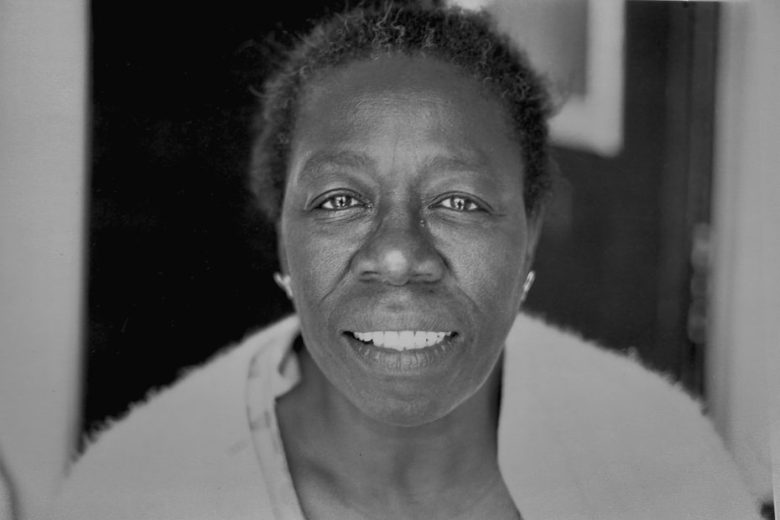 Black and white photo of an older woman with short hair, wearing a light top and earrings, looking directly at the camera with a neutral expression.