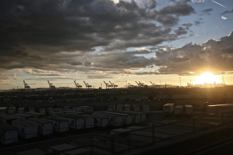 Shipping containers and cranes at a dockyard are silhouetted against a cloudy sky at sunset, with the sun low on the horizon.