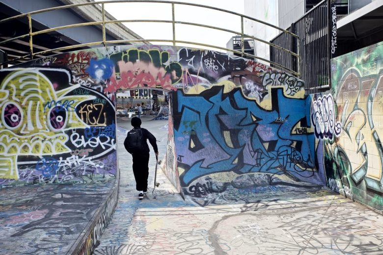 A person with a backpack walks through a graffiti-covered tunnel in an urban setting, surrounded by colorful street art and tags.