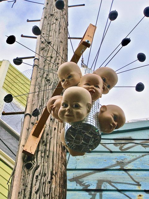 A cluster of doll heads is mounted on a wooden pole, surrounded by metal rods with black ends, against a background of buildings and sky.