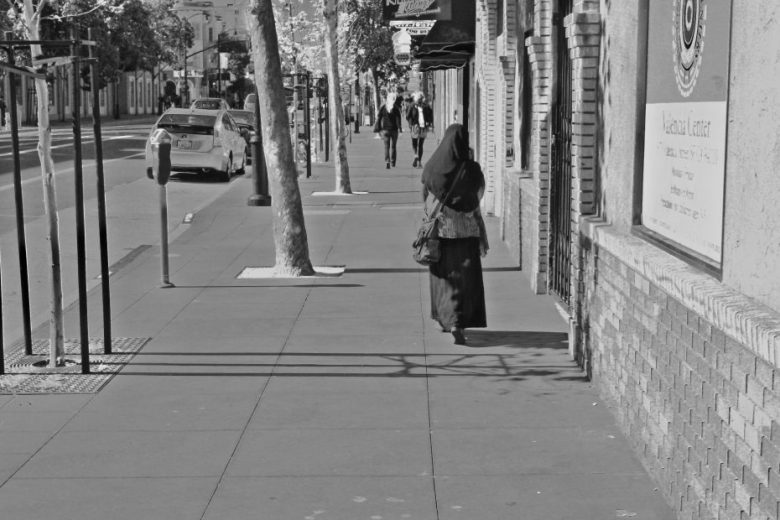 A woman wearing a headscarf and long dress walks down a city sidewalk in daylight; a car and pedestrians are visible further down the street.