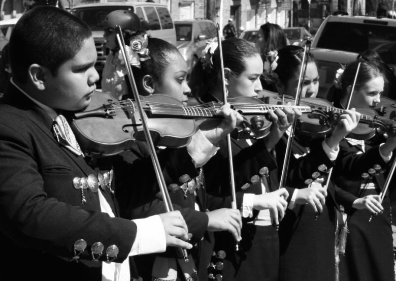A group of musicians, dressed in traditional attire, stand outdoors in a line playing violins. The image is in black and white.