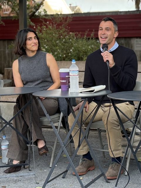 Two people sit at a table outdoors; the man on the right is speaking into a microphone while the woman on the left listens. Drinks and notebooks are on the table.