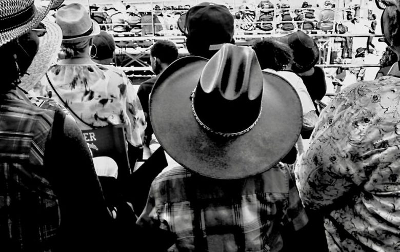 A group of people viewed from behind, many wearing hats, with a focus on one person in a cowboy hat, facing bleachers in an outdoor setting.